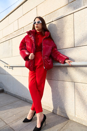 High fashion photo of a young brunette woman in red glossy down jacket, and red pants. Urban street...