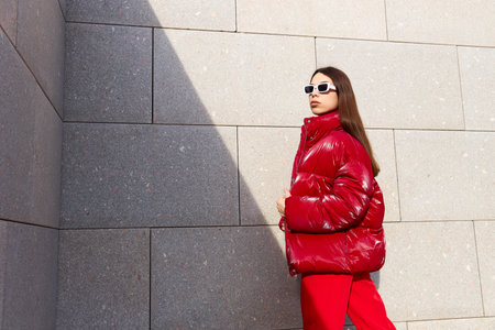 High fashion photo of a young brunette woman in red glossy down jacket, and red pants. Urban street fashion. Monochrome red outfit, white sunglassesの写真素材