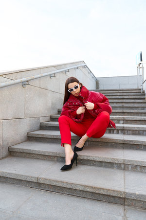 High fashion photo of a young brunette woman in red glossy down jacket, and red pants. Urban street fashion. Monochrome red outfit, trendy white sunglassesの写真素材