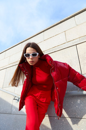 High fashion photo of a young brunette woman in red glossy down jacket, and red pants. Urban street...