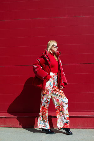 High fashion photo of a young blond woman in red glossy down jacket, poloneck, and pants. Monochrome trendy red outfit. Urban street fashionの写真素材
