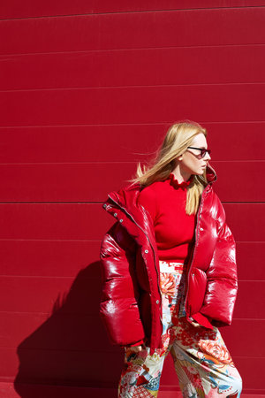 High fashion photo of a young blond woman in red glossy down jacket, poloneck, and pants. Monochrome trendy red outfit. Urban street fashionの写真素材