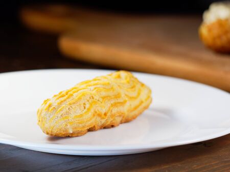 One eclair on a white plate, on a dark wooden background. Homemade baking.