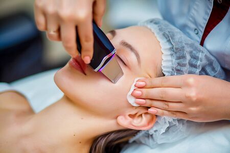Woman receiving ultrasonic facial exfoliation at cosmetology salon. Procedure clearing clogged pores, ultrasonic treatment for skin rejuvenation, beautician uses modern apparatus for refreshingの写真素材