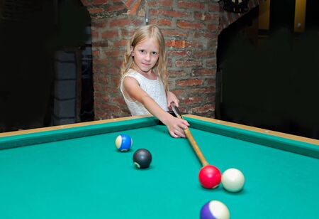 Little beautiful blond girl learns to play billiards, pool, snooker, Russian pyramid in kid's club.