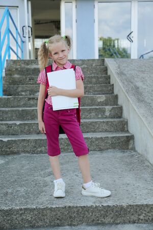 Back to school. Beautiful blond schoolgirl with red backpack and books outside the primary school , education conceptの写真素材