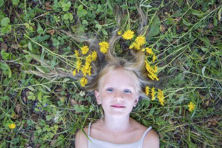 Fashion art portrait young little blonde girl, hair waves lie on the green grass with yellow flowers. Top view, outdoors.の写真素材
