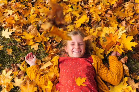 Happy little girl playing with fallen yellow leaves in autumn park. Girl with blonde hair lying on...