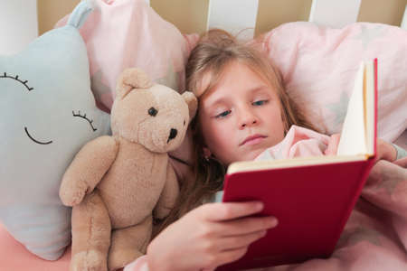 Little blonde girl readingbook . Girl lying on white bed and reading book with her toy bear.