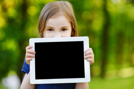 Happy little girl holding tablet PC outdoors in summer park on beautiful sunny dayの写真素材