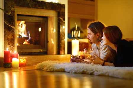 Happy couple having drinks by a fireplace in a cozy dark living room on Christmas eveの写真素材
