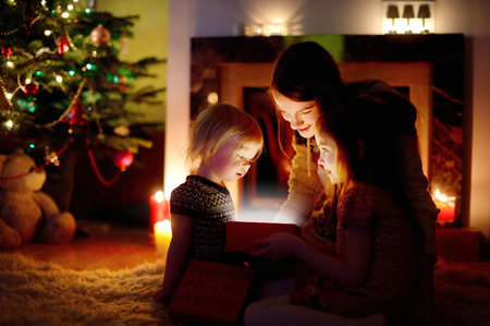 Young mother and her two little daughters opening a magical Christmas gift by a Christmas tree in cozy living room in winterの写真素材