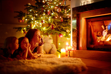 Young mother and her two little daughters sitting by a fireplace in a cozy dark living room on Christmas eveの写真素材