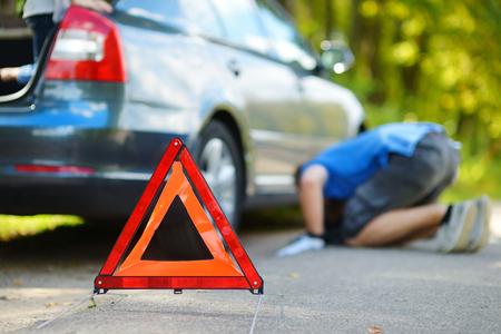 Red warning triangle sign on the road with a man checking his broken car in backgroundの写真素材