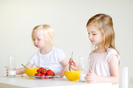 Two cute little sisters eating cereal with strawberries and drinking milk in white kitchenの写真素材