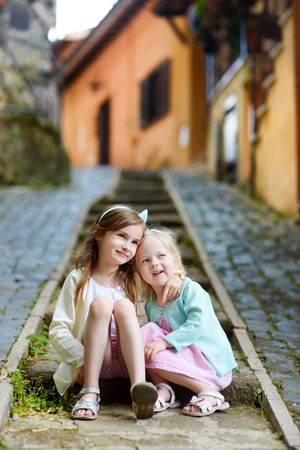 Two adorable little sisters laughing and hugging each other on warm and sunny summer day in italian townの写真素材
