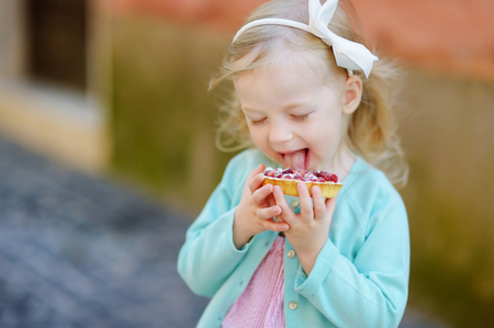 Adorable little girl eating fresh sweet strawberry cake outdoors on warm and sunny summer day in Italyの写真素材