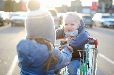 Two adorable sisters playing at parking lot with a shopping cart at cold autumn dayの写真素材