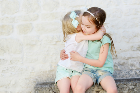 Two adorable little sisters laughing and hugging each other on warm and sunny summer day in italian townの写真素材