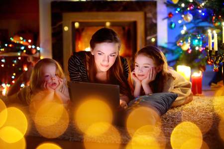 Young mother and her daughters using a tablet pc by a fireplace on warm Christmas eveningの写真素材