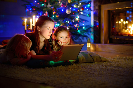Young mother and her daughters using a tablet pc by a fireplace on warm Christmas eveningの写真素材