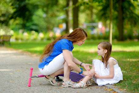 Mother comforting her daughter after she fell while riding her scooter at summer parkの写真素材