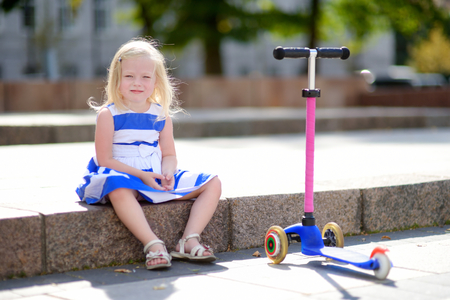 Adorable little girl wearing beautiful dress riding her scooter in a summer parkの写真素材
