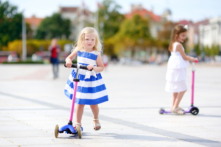 Two adorable little sisters wearing beautiful dresses riding their scooters in a summer parkの写真素材