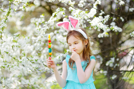 Adorable little girl wearing bunny ears eating colorful gum candies on a stick in blooming cherry garden on Easter dayの写真素材