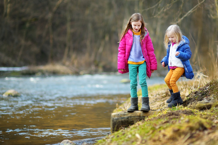 Two cute little sisters playing with a stick by a river on warm spring dayの写真素材