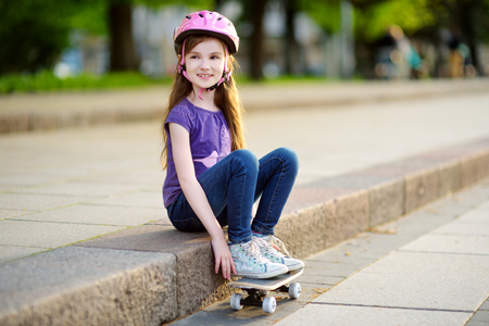 Cute little preteen girl wearing helmet sitting on a skateboard in beautiful summer parkの写真素材