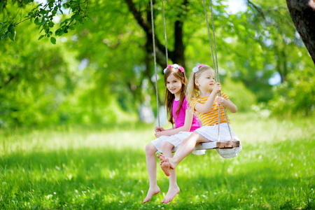 Two cute little sisters having fun on a swing together in beautiful summer garden on warm and sunny day outdoorsの写真素材