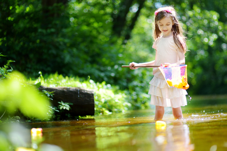 Cute little girl playing in a river catching rubber ducks with her scoop-net on warm and sunny summer dayの写真素材