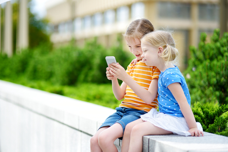 Two cute little sisters playing outdoor mobile game on their smart phones. Kids catching virtual pocket monsters. Modern addictive multiplayer location-based games.の写真素材