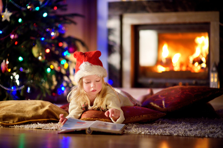 Adorable little girl reading a story book under a Christmas tree on Christmas eve at homeの写真素材