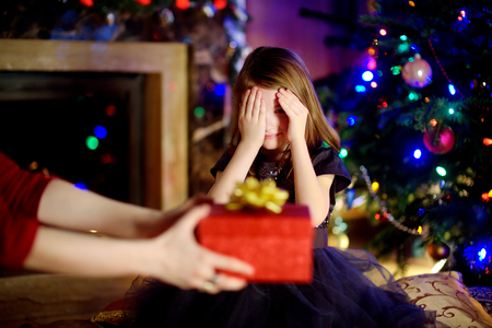 Happy little girl getting a Christmas gift from her mommy by a fireplace on Christmas eveの写真素材