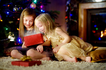 Happy little sisters opening magical Christmas gift by a fireplace in a cozy dark living room on Xmas eveの写真素材