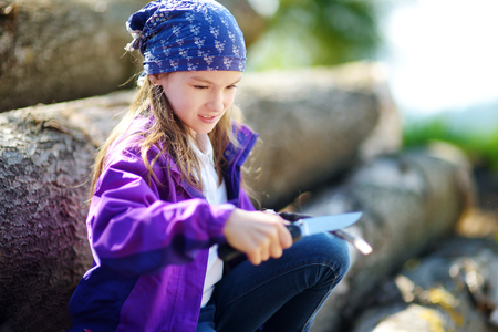 Cute little girl sitting on tree logs using a pocket knife to whittle a hiking stick. Child using a carving knife.の写真素材