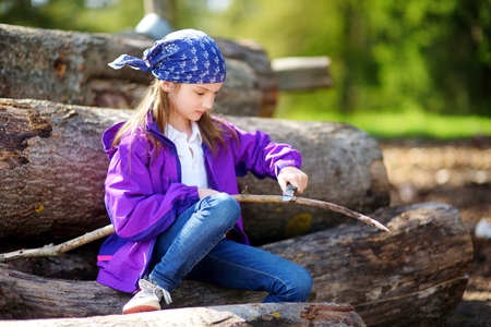 Cute little girl sitting on tree logs using a pocket knife to whittle a hiking stick. Child using a carving knife.の写真素材