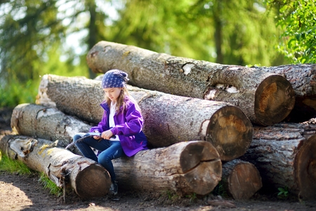 Cute little girl sitting on tree logs using a pocket knife to whittle a hiking stick. Child using a carving knife.の写真素材
