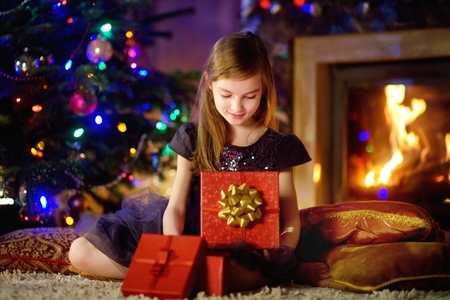 Happy girl opening Christmas gifts by a fireplace in a cozy dark living room on Xmas eveの写真素材