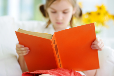 Adorable little girl reading a book in white living room on beautiful summer dayの写真素材