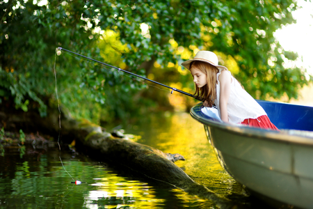 Cute little girl having fun in a boat by a river at beautiful summer evening. Child fishing with a fishing rod on warm and sunny day.の写真素材