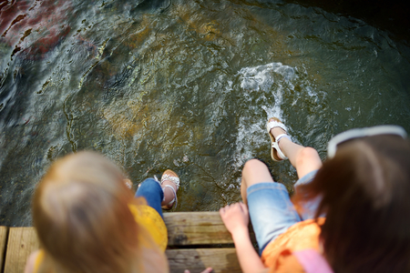 Two cute little girls sitting on a wooden platform by the river or lake dipping their feet in the water on warm summer dayの写真素材