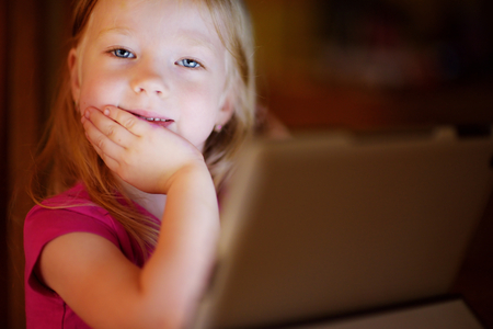 Adorable little girl playing with a digital tablet in a dark room. Child in an elementary school. Education and learning for kids.の写真素材