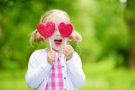 Cute little girl eating huge heart-shaped lollipop outdoors on beautiful summer day. Child with lolly candy outdoors.の写真素材