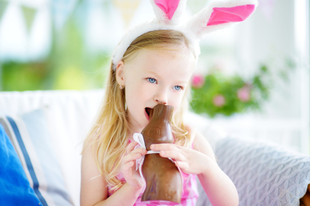 Cute little girl wearing bunny ears eating chocolate Easter rabbit. Kid playing egg hunt on Easter. Adorable child celebrate Easter at home.の写真素材