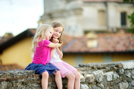 Two adorable little sisters laughing and hugging on warm and sunny summer day in italian townの写真素材