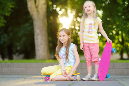 Two pretty little girls learning to skateboard on beautiful summer day in a park. Children enjoying skateboarding ride outdoors. Summer activities for kids.の写真素材