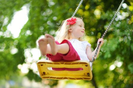 Cute little girl having fun on a playground outdoors on warm summer day. Cute kid swinging outdoors. Summer outdoor leisure for kids.の写真素材